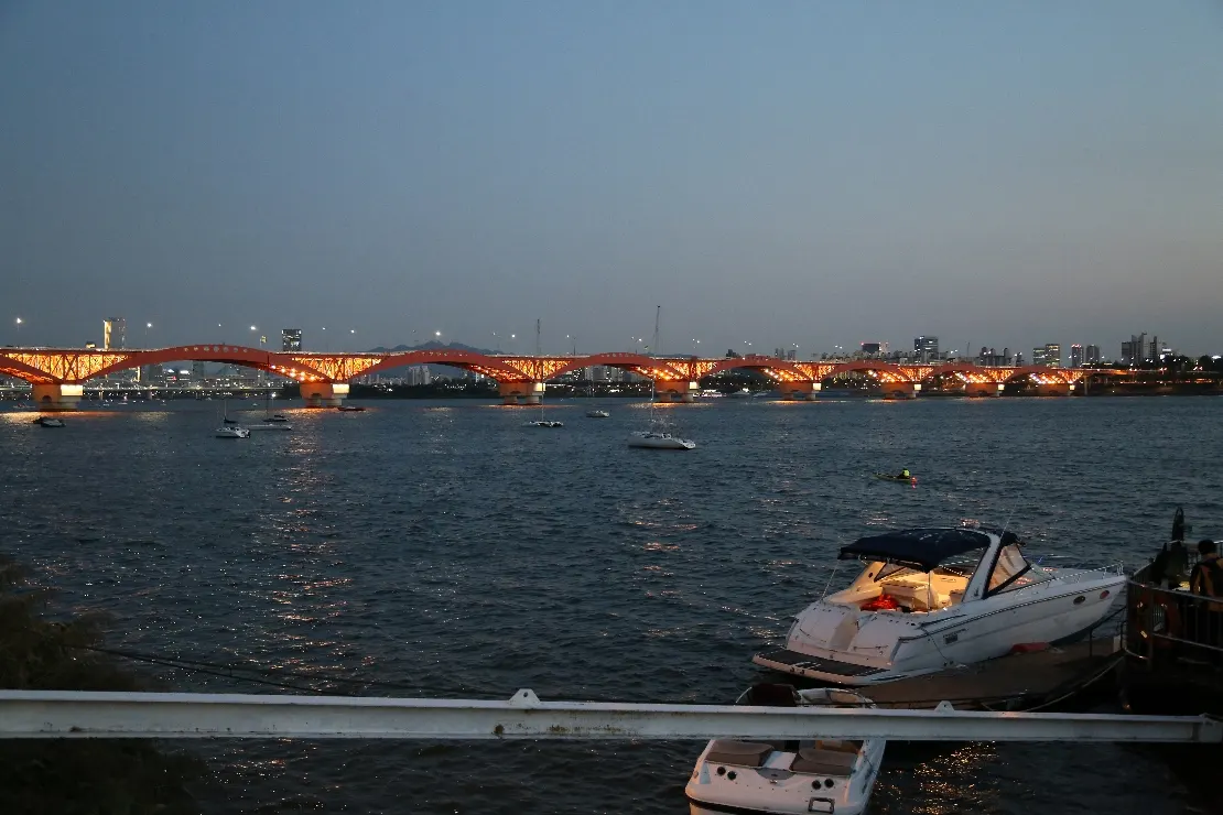 An evening river view from Mangwon Hangang Park, with bridge lights and boats by the bank.